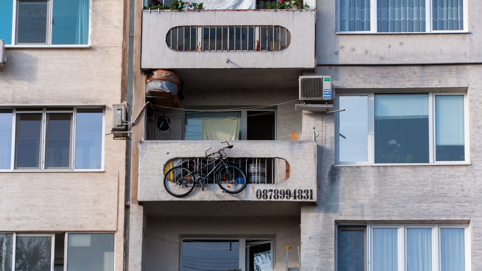 Urban scene of a concrete apartment building with a bicycle on the balcony and visible air conditioning unit.