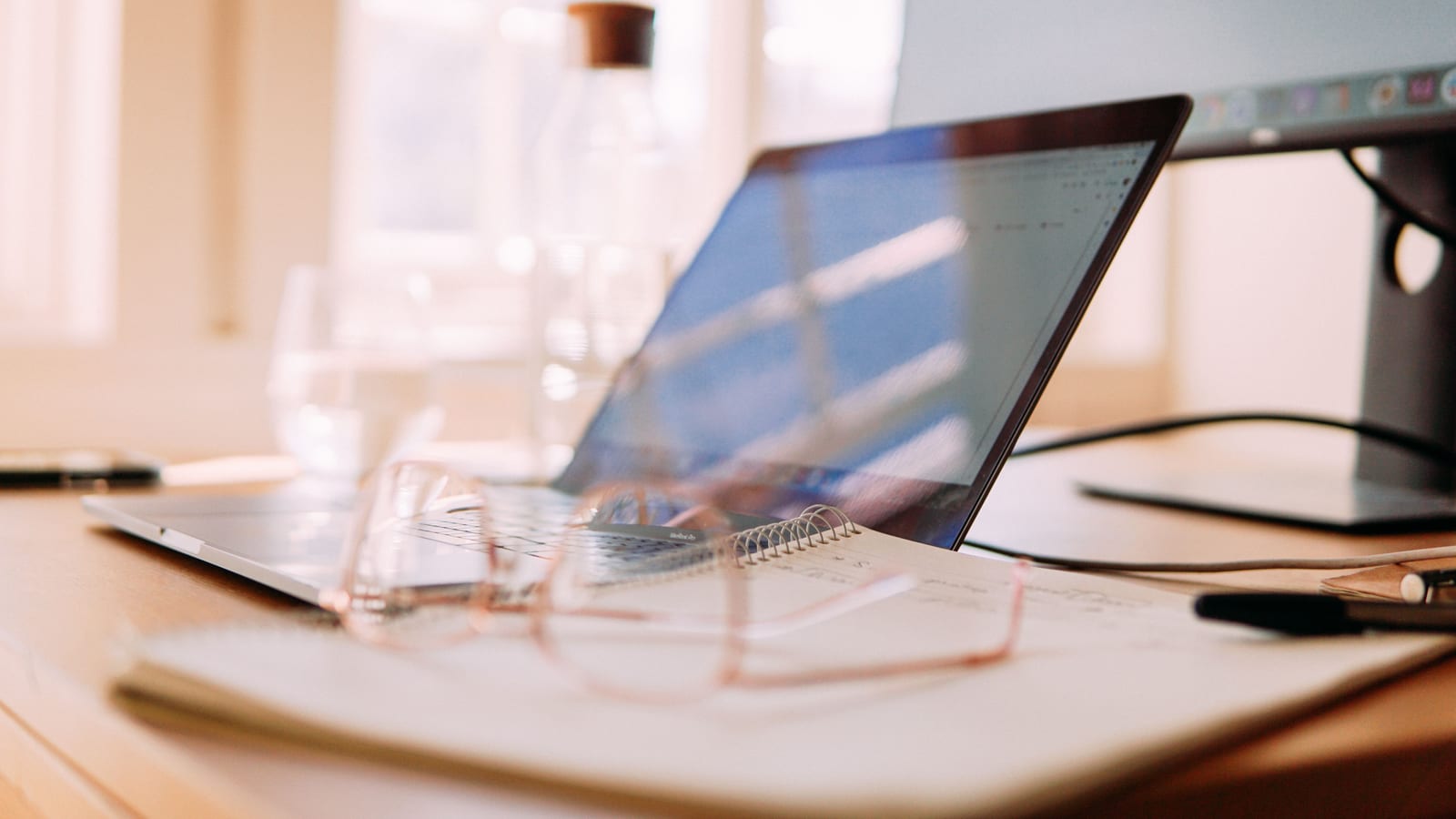 A well-lit home office setup featuring a laptop, monitor, and glasses, ideal for remote work.